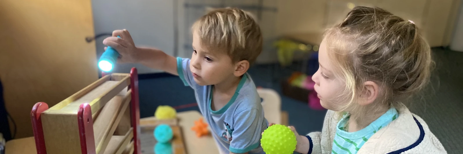 Two children using flashlights to explore a provocation featuring mirrors, an inclined ramp, and balls, sparking curiosity and investigation.