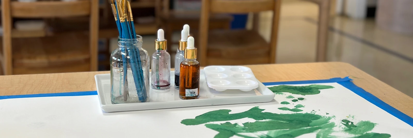 wide shot of a classroom with a table in the foreground set up with paintbrushes and watercolors. The paper on the table displays forest green marks left by one of the students. In the background, the communication space features a table, wonder questions, and line tools, inviting exploration and dialogue.