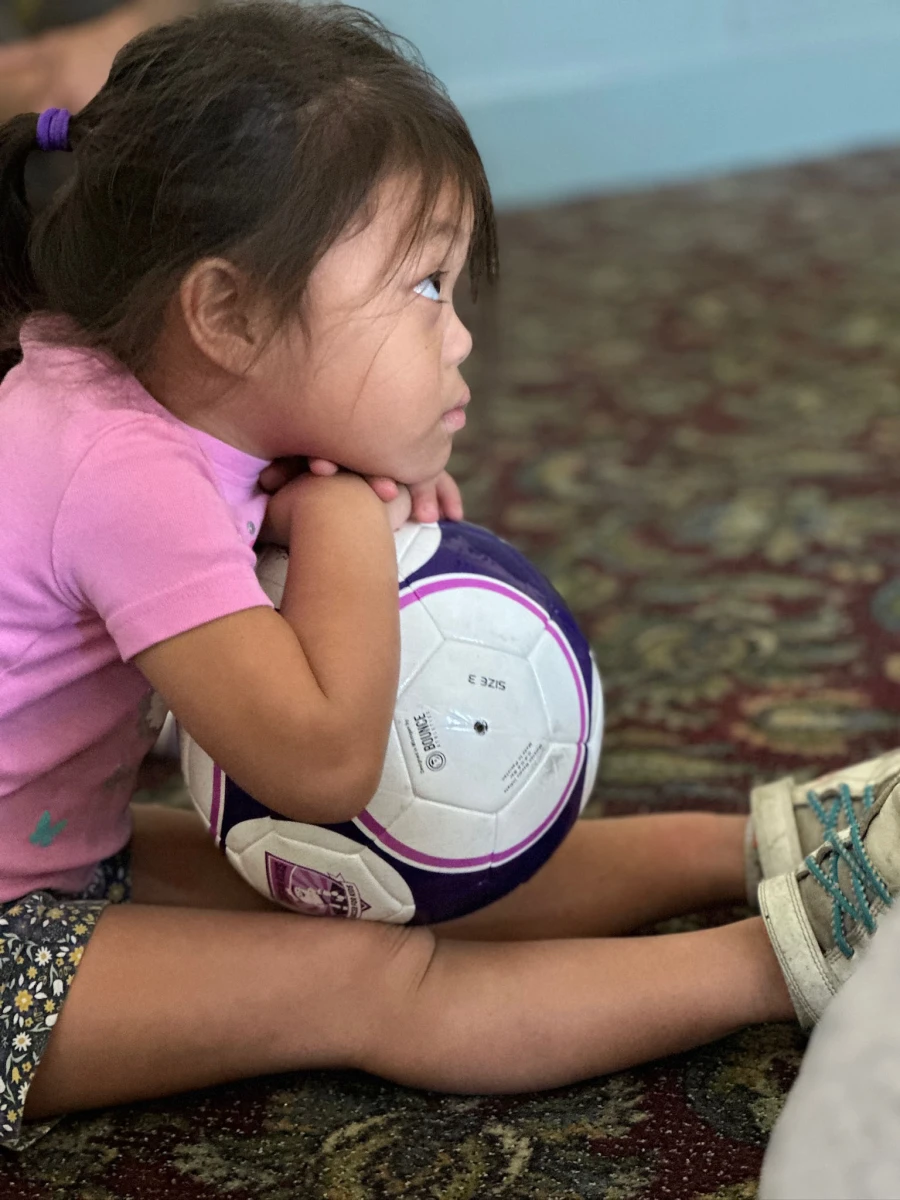 A child sitting with their legs outstretched, eagerly holding a soccer ball, awaiting instructions from the coach with anticipation.