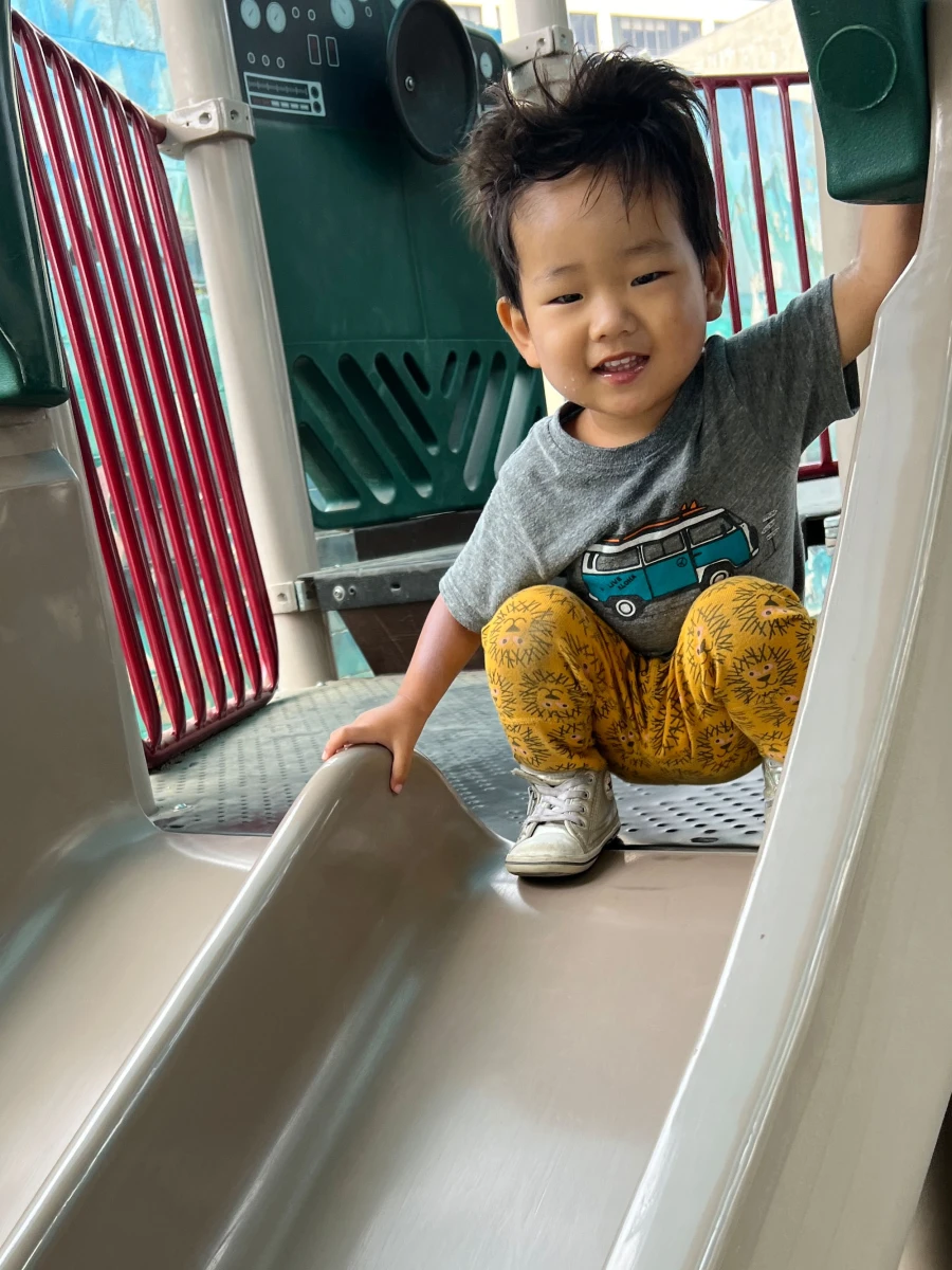 A toddler happily perched at the top of the slide, eagerly getting ready to take off with excitement.