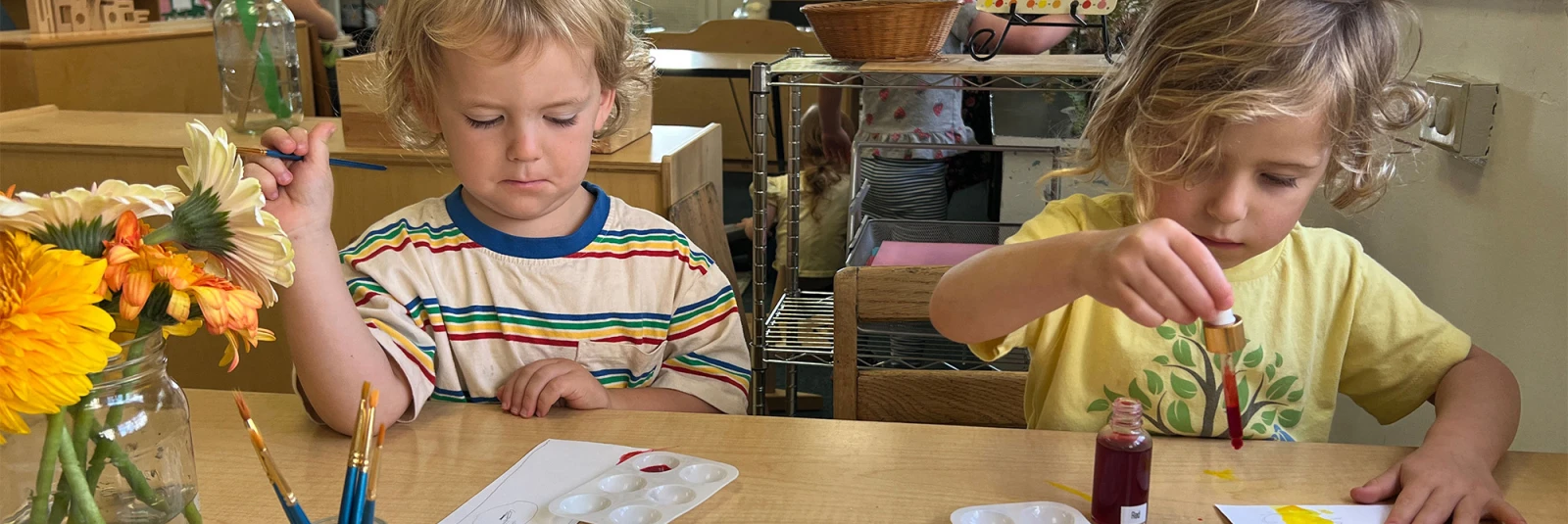 A wide shot of two children seated at a table in a classroom filled with natural light, working with watercolors to create artwork inspired by yellow, cream, and orange flowers.