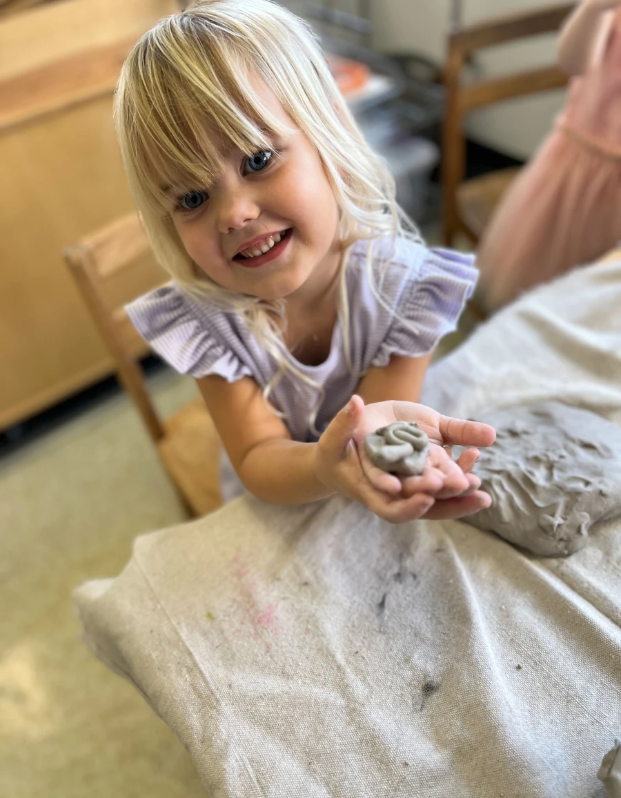 A child with a big smile, proudly holding a clay creation as if presenting a very special treasure.
