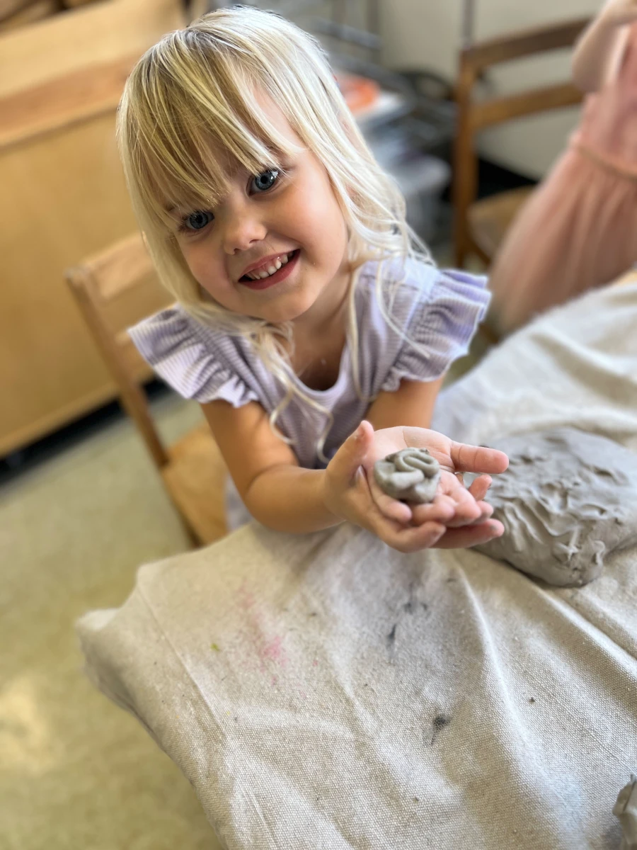 A child with a big smile, proudly holding a clay creation as if presenting a very special treasure.