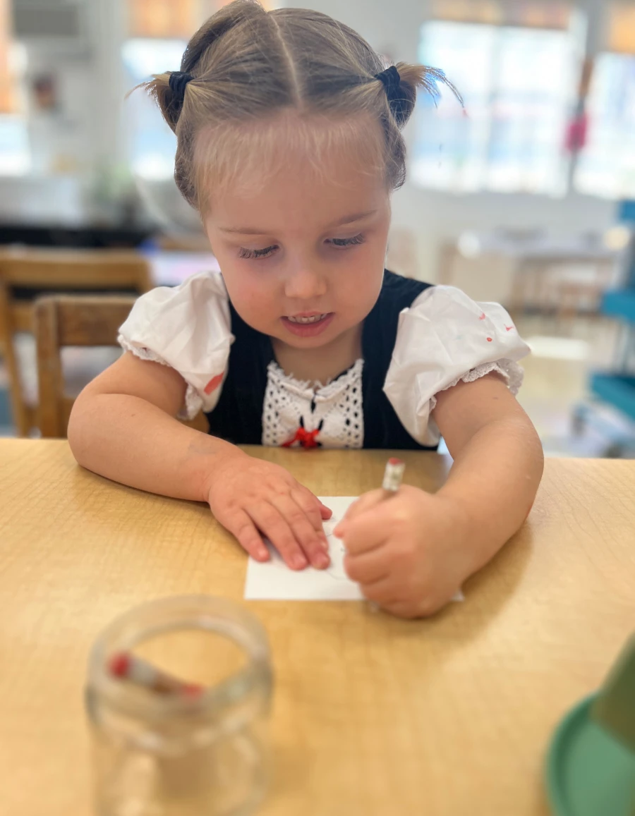 A young child seated at a table, holding a pencil, deeply focused on creating a self-portrait.