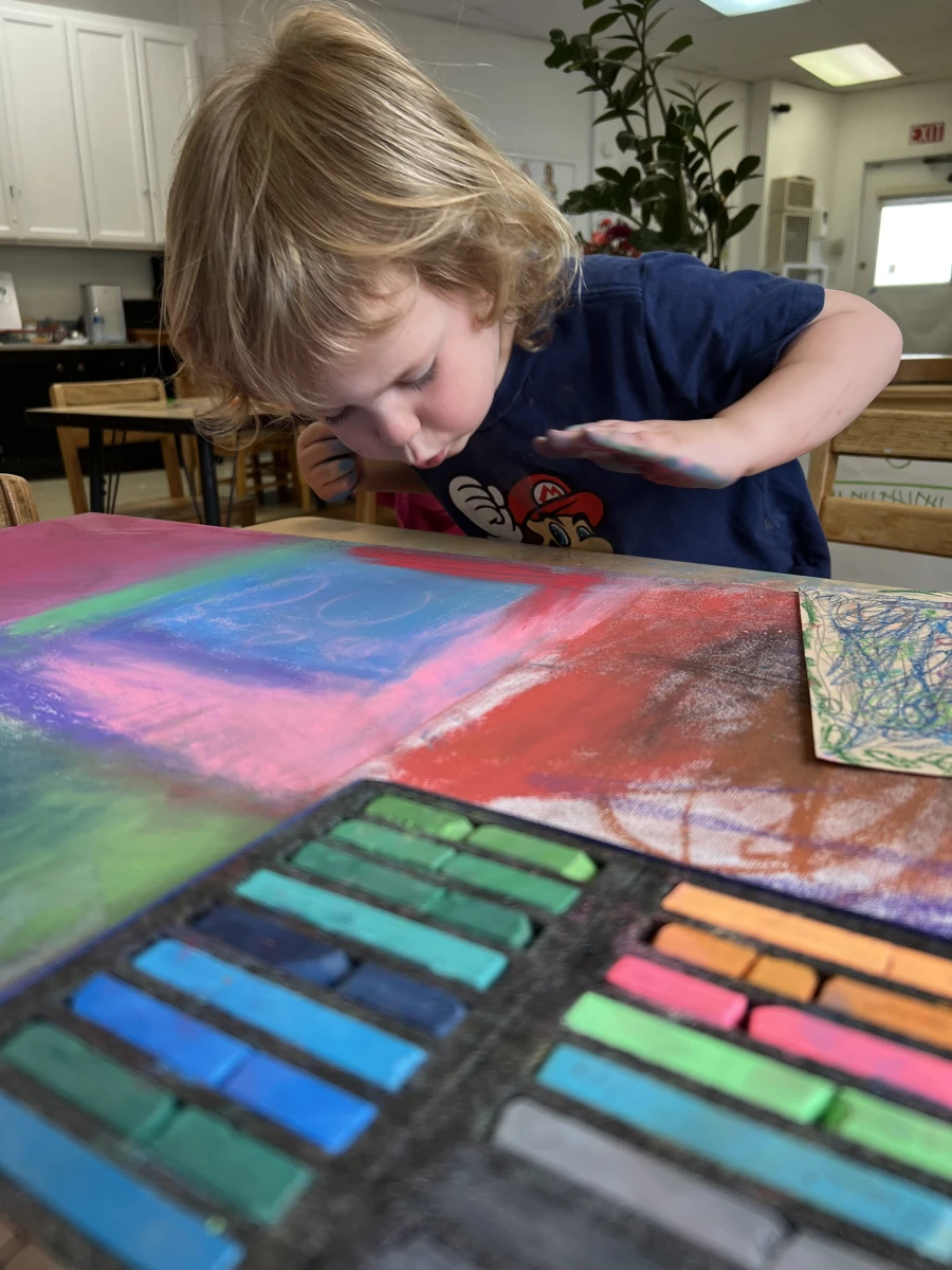 In the foreground, a brightly colored pastel artwork and a box of pastels take center stage. In the background, a child with blue chalk on his hands carefully blows away the excess dust, revealing the masterpiece.