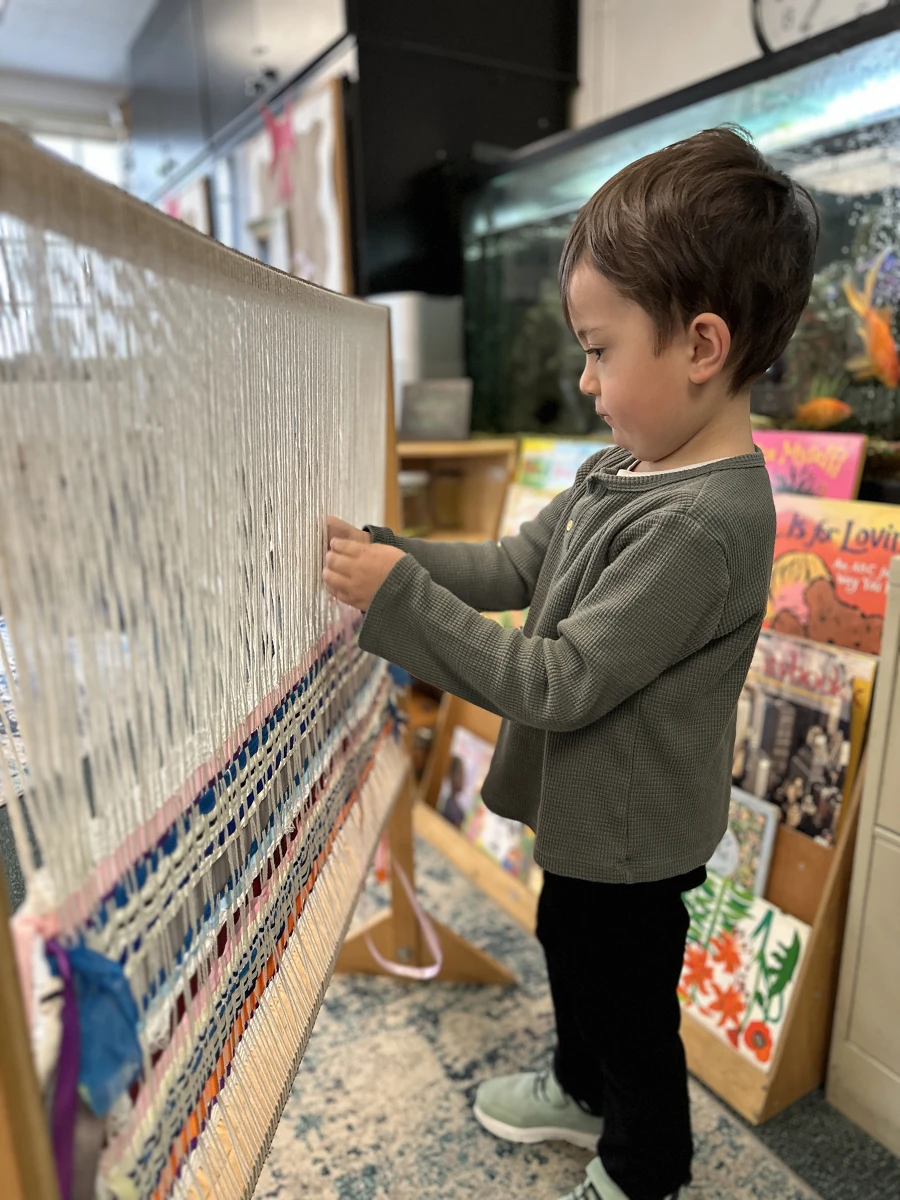 A child carefully working on a large loom, focused on weaving. In the background, a shelf filled with books and a fish tank add to the cozy, creative atmosphere.