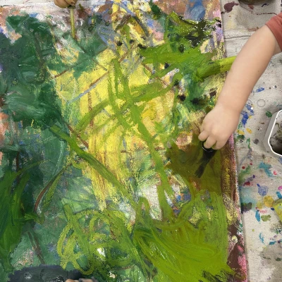 A top-down view of a canvas filled with a variety of greens, blues, and yellows blending together. Three small hands are visible, holding tempera sticks and eyedroppers as they continue their creative work.
