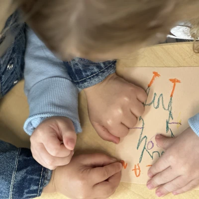 A top-down view of two children working on a note, showing only their hands, the paper, and the colorful pastels they are using.